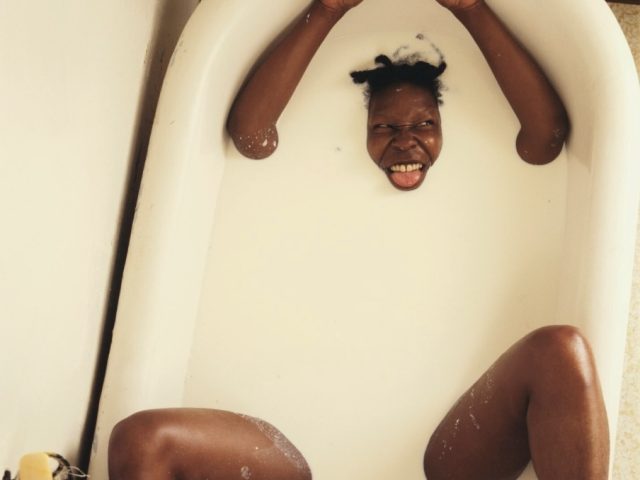 Person submerged in milky bathwater with head, arms, and legs above the surface, grinning playfully and sticking out tongue.